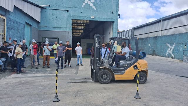 Trabalhador opera uma empilhadeira entre cones, observado pela turma do treinamento, em frente a um galpão.